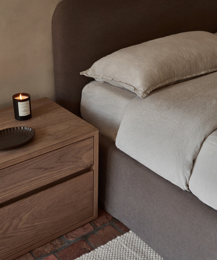 A neatly made bed featuring the Dusk Linen Flat Sheet in light beige with a brown upholstered headboard sits beside a wooden nightstand holding a lit candle and dark dish, atop a brick floor with a woven rug visible.