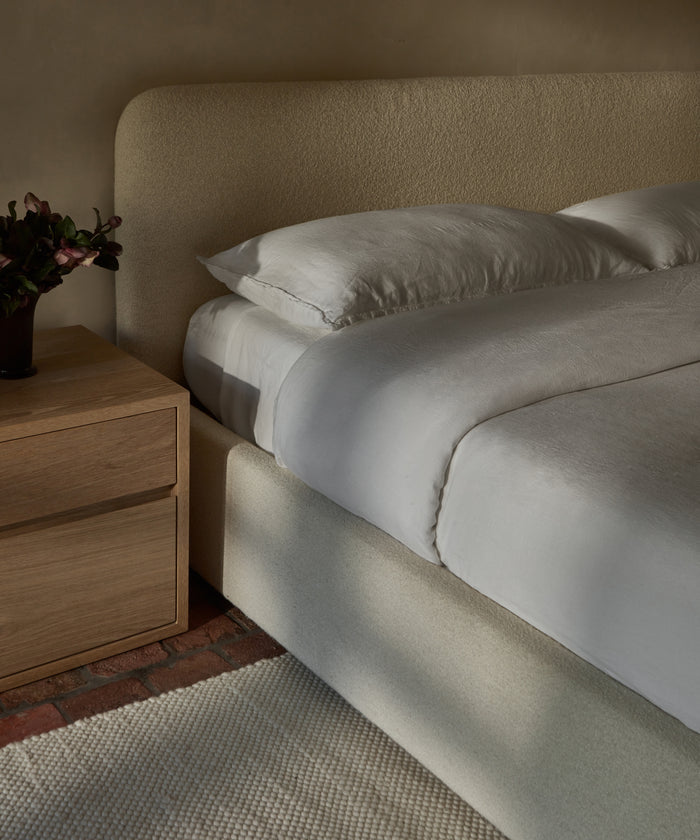 A neatly made bed with the Dusk Linen Flat Sheet and light beige bedding, featuring a curved upholstered headboard next to a wooden nightstand with a dark vase of flowers—set atop a textured cream rug and brick flooring.