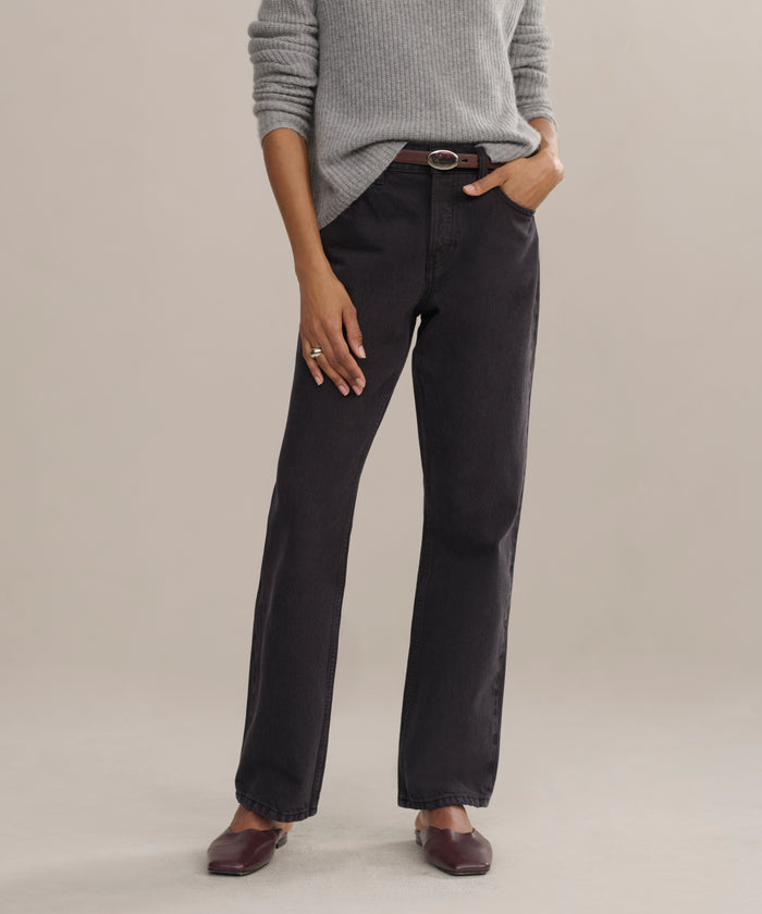 A person wears the Easy Straight Crop luxury jeans in a straight cropped leg and mid-rise fit, paired with a light gray sweater, brown belt, and brown shoes, standing with one hand in their pocket against a plain background.