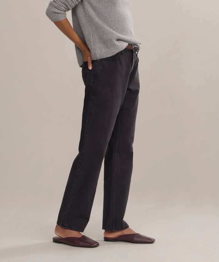 A person stands sideways with one hand on their hip, wearing a gray sweater and the Easy Straight Crop luxury jeans in a mid rise, straight cropped fit, paired with brown flat shoes against a neutral background. Their face is not visible.
