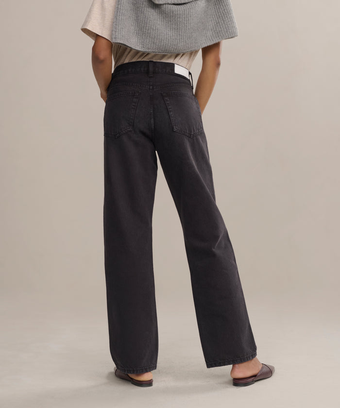 A person stands with their back to the camera, wearing the Easy Straight Crop luxury jeans with a mid-rise fit and wide-leg cut, paired with a beige top, gray sweater over their shoulders, brown flats, and set against a neutral background.