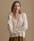 Woman wearing an ivory cardigan with brown shorts against a beige wall.
