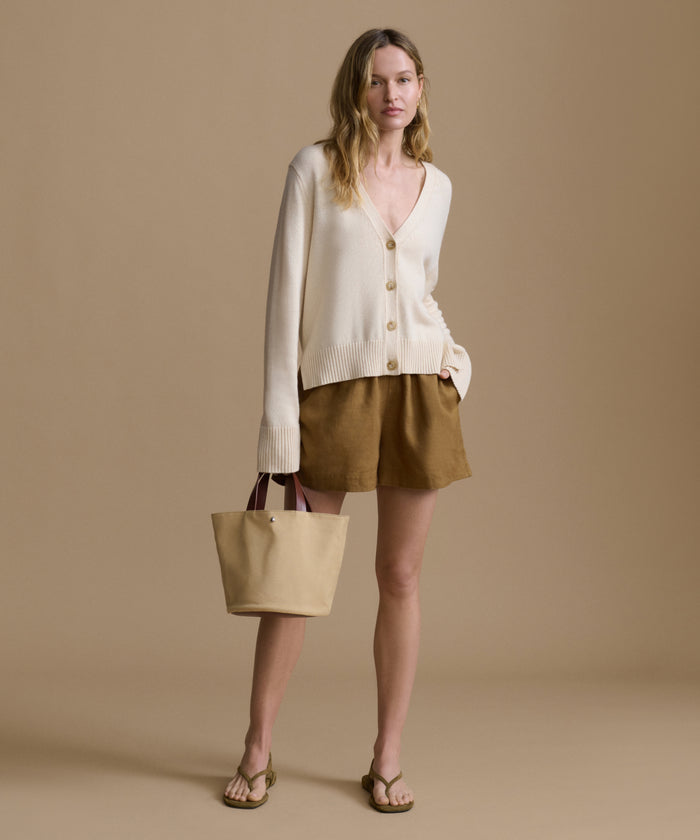 Woman wearing an ivory cardigan with brown shorts carrying a handbag against a beige wall.