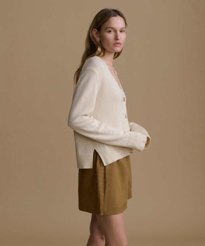 Woman wearing an ivory cardigan with brown shorts against a beige wall.