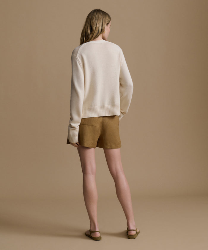 Woman wearing an ivory cardigan with brown shorts against a beige wall.