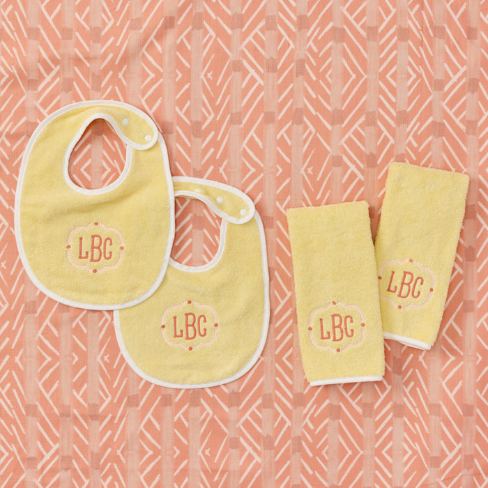 Flat lay of two yellow baby bibs and two burp cloths with pink LBC monogram on orange background.