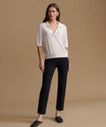 A woman stands against a warm beige studio background wearing a cream short-sleeve knit polo-style top and black cropped trousers with dark loafers.
