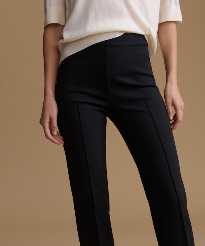 A model wearing black high-waisted trousers with front pleats and a cream-colored short-sleeve top, set against a solid warm beige background.