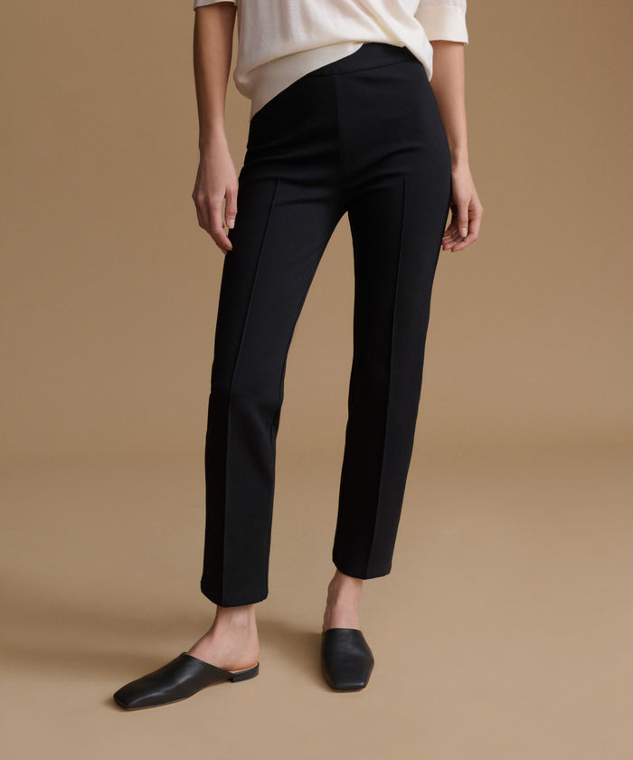 A model wearing black straight-leg ankle-length trousers, styled with a cream top and black mule flats, set against a solid beige studio background.