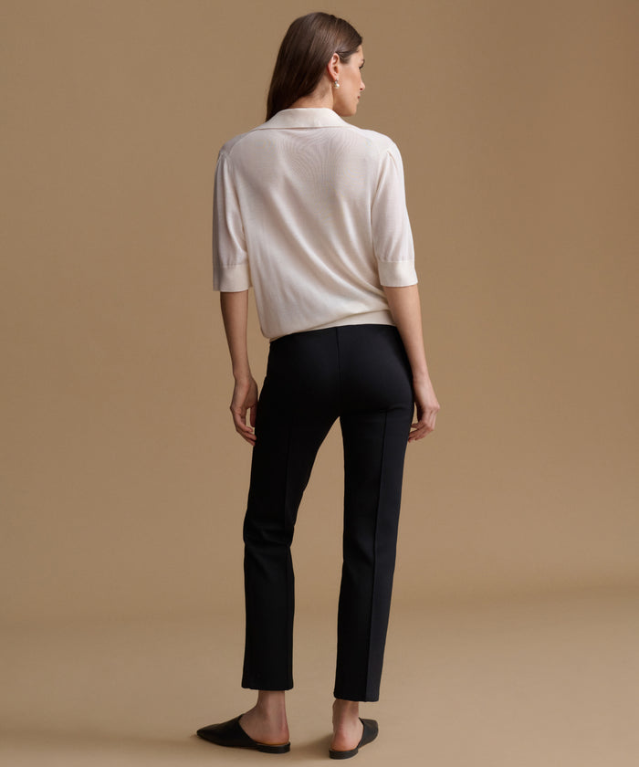 A woman shown from the back wearing a cream-colored knit polo shirt and black trousers, standing against a solid beige studio background.