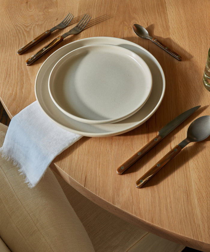 A wooden dining table set with two stacked cream-colored plates, a Frayed Linen Napkin, a fork and spoon above the plates, a knife and spoon to the right, and part of a beige chair visible in the lower left corner.