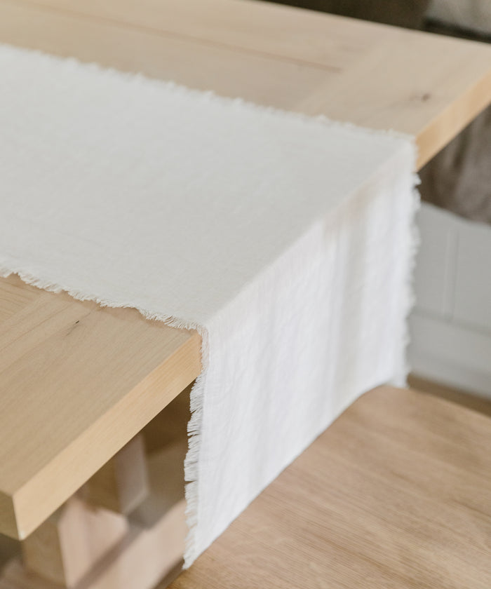 A light wood table is adorned with the Frayed Linen Runner in ivory, featuring a soft texture and delicate fringes that drape gracefully over the edge.