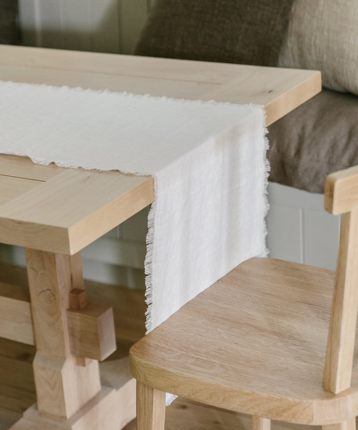 A light wooden dining table features a Frayed Linen Runner with fringed edges draped over it. A matching wooden chair sits beside the table, complemented by neutral cushions in the background for a calming vibe.