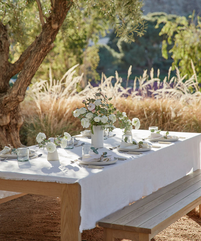 A wooden outdoor table draped in the Frayed Linen Tablecloth is set with white dishes, glasses, and white flowers. It sits beneath a tree amid tall grass, creating a sunlit natural setting.