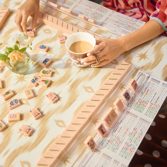 A top-down lifestyle shot of a Mahjong game in progress. Hands are visible moving wooden tiles and holding a white mug over a tan and cream ikat patterned tablecloth. The table is set with Mahjong racks, scattered tiles, a scoring card, and a small vase of flowers.