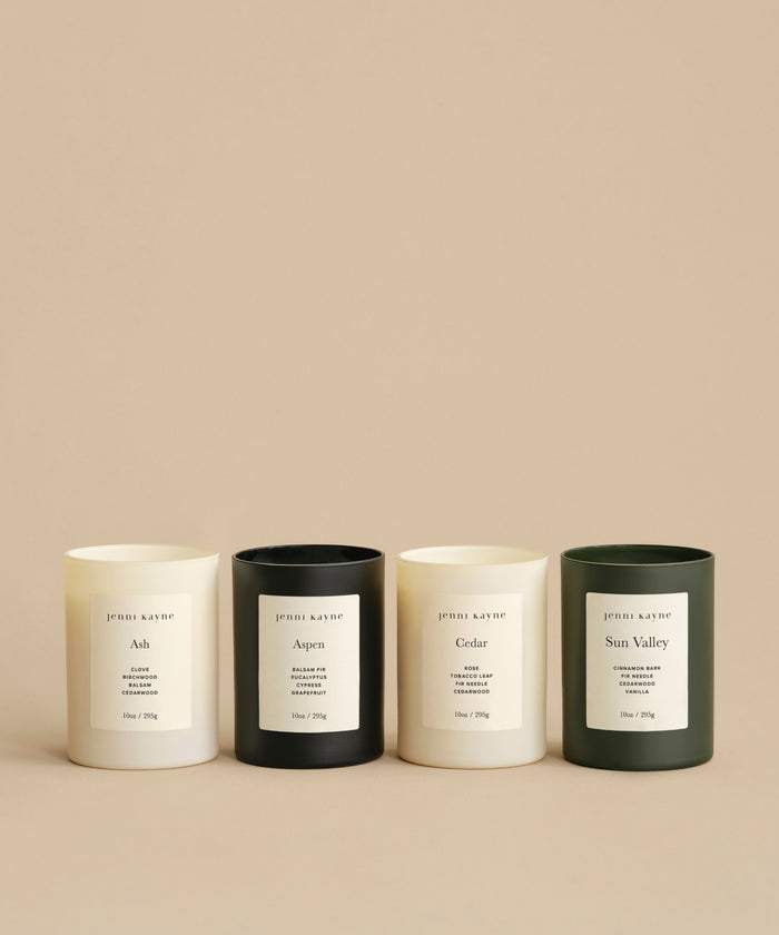 The Glass Candle Gift Set includes four scented candles in minimalist jars—two cream, one black, and one dark green—each labeled with the scent name: Ash, Aspen, Cedar, or Sun Valley. Perfectly lined up against a beige background.