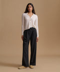 A woman stands against a warm beige studio background wearing a light cream long-sleeve button-front top, black wide-leg pants, and tan slip-on shoes.