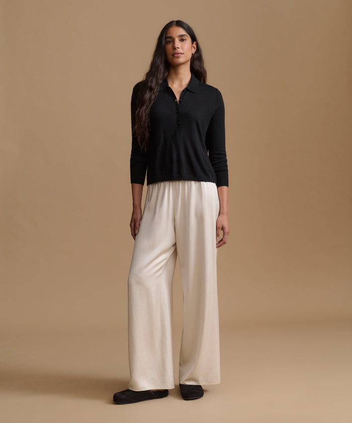 A woman stands against a warm beige studio background wearing a black long-sleeve polo-style top and wide-leg cream pants with black shoes.