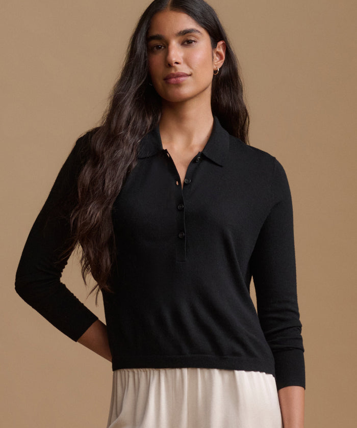 A woman modeling a black long-sleeve polo knit top with a buttoned placket, paired with a light cream skirt, against a solid warm beige studio background.