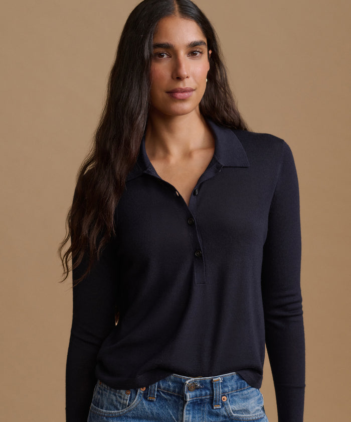 A woman modeling a dark navy long-sleeve polo shirt with a buttoned collar, paired with blue jeans, against a solid warm beige background.