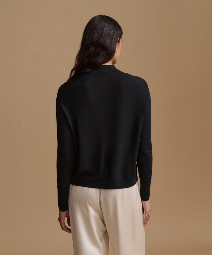 A woman viewed from the back wearing a black long-sleeve mock neck knit top and cream-colored trousers, set against a solid warm beige studio background.