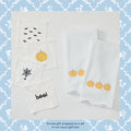 A set of white Halloween-themed baby textiles, including two ribbed square cloths with embroidered pumpkins and three smaller square cloths with embroidered bats, a spider web, and the word 'boof!' in black, displayed on a white background with a blue floral border.