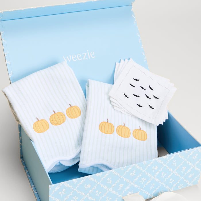 A baby gift set in a light blue box, featuring three folded white striped baby towels with embroidered orange pumpkins, a stack of white baby napkins with black bat silhouettes, and the brand name 'weezie' printed inside the box.