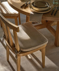 A wooden dining chair with a beige Hampton Chair Cushion featuring a removable cover sits by a round wooden table set with plates, bowls, napkin, and cutlery. Sunlight casts warm shadows on the light wood floor.