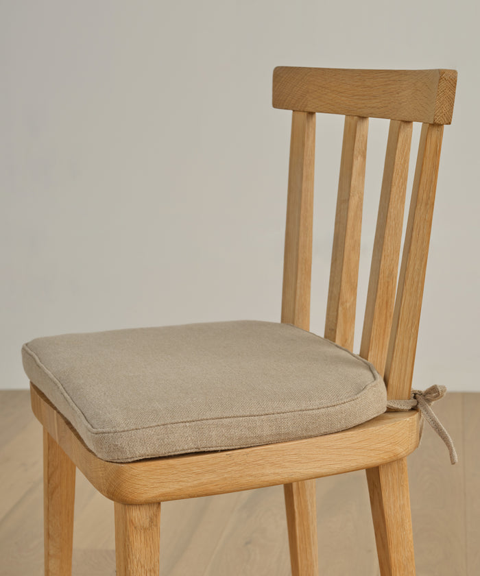 A wooden chair with a beige Hampton Chair Cushion made of Belgian linen, featuring a removable cover tied to the seat, sits on a wooden floor against a plain light wall.