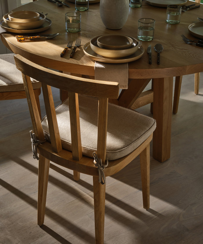 A wooden dining chair with a Hampton Chair Cushion in beige Belgian linen is placed by a round wooden table set with plates, glasses, and cutlery in natural light. The table setting is simple and elegant.