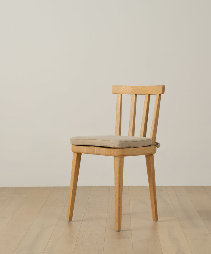 The Hampton Chair Cushion, upholstered in beige Belgian linen with a removable cover, rests on a simple wooden chair placed on a light wood floor against an off-white wall.