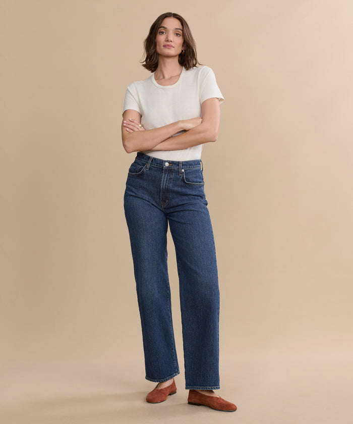 A person stands against a beige background wearing a white short-sleeve t-shirt, the Harper Jean in organic cotton with a wide-leg silhouette, and brown shoes, arms crossed and looking at the camera.