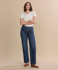 A person stands against a beige background wearing a white short-sleeve t-shirt, the Harper Jean in organic cotton with a wide-leg silhouette, and brown shoes, arms crossed and looking at the camera.