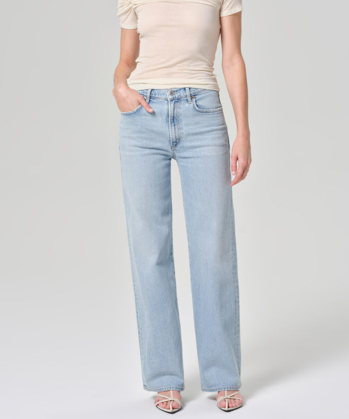 Wearing a beige short-sleeve top, Harper Jean in light blue organic cotton with a wide-leg silhouette, and white sandals, this person stands against a plain light background with one hand in their pocket.