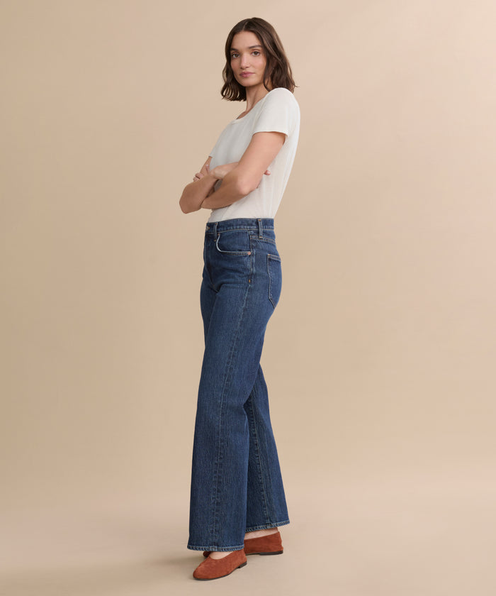 A person stands sideways with arms crossed, wearing a white short-sleeve shirt, the Harper Jean in an organic cotton wide-leg silhouette, and brown shoes against a plain beige background.