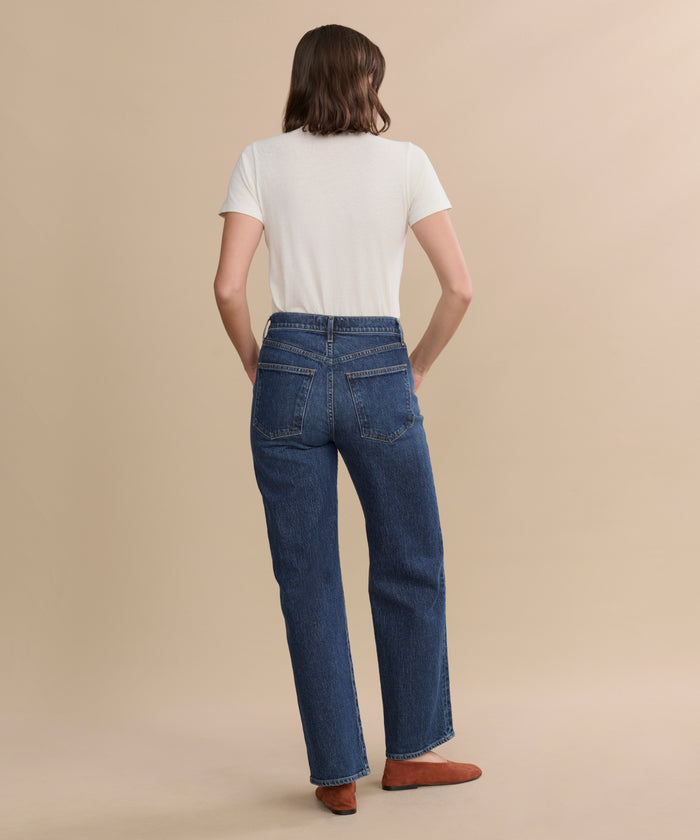 A person with shoulder-length brown hair stands facing away, wearing a white short-sleeve t-shirt, brown shoes, and the Harper Jean—relaxed mid-rise jeans with a wide-leg silhouette—against a plain beige background.