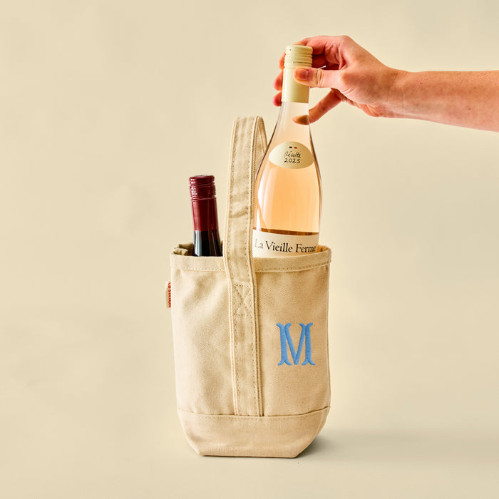 A hand placing a bottle of rosé wine into a cream canvas tote bag that features a blue embroidered 'M' monogram.