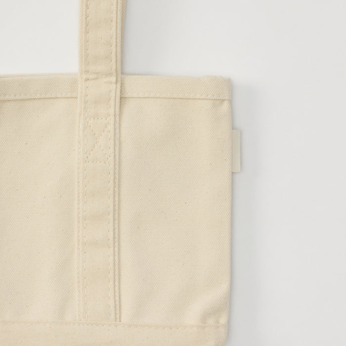 A close-up of a plain cream canvas tote bag showing the stitched handle strap and a small tag on the side seam.