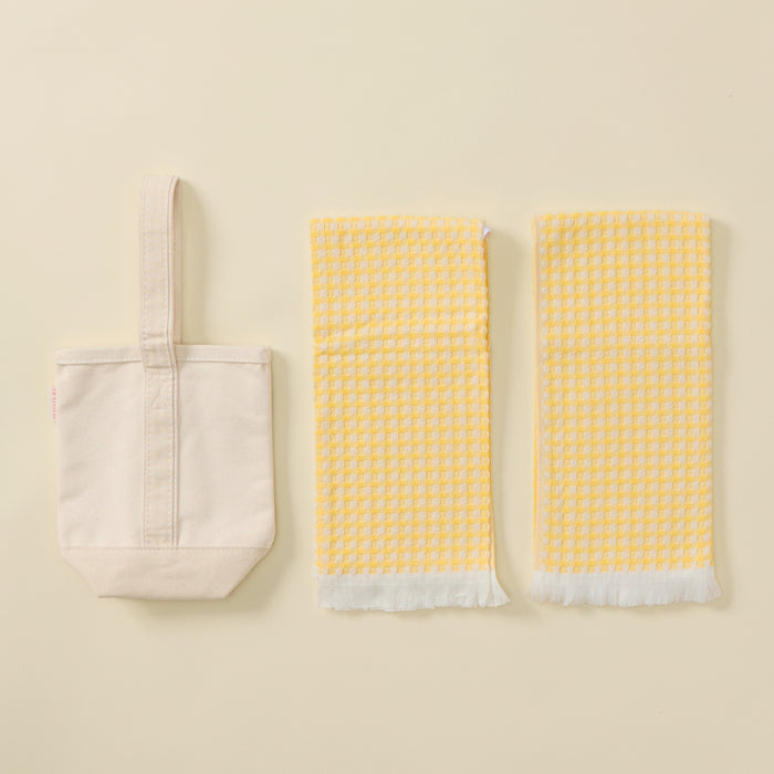 A small off-white fabric wine bag on the left, and two rectangular yellow-and-white checkered towels with white fringed edges on the right.