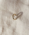 The Huggie, a small 14K gold hoop earring, sits on textured off-white fabric and casts a subtle shadow—ideal for those who love minimalist jewelry.