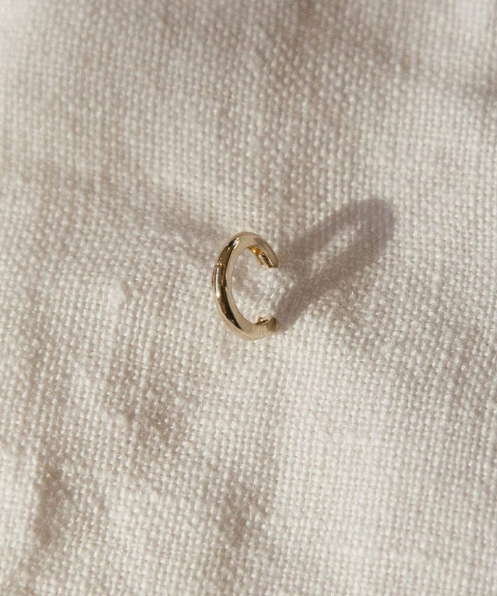 The Huggie, a small 14K gold hoop earring, sits on textured off-white fabric and casts a subtle shadow—ideal for those who love minimalist jewelry.