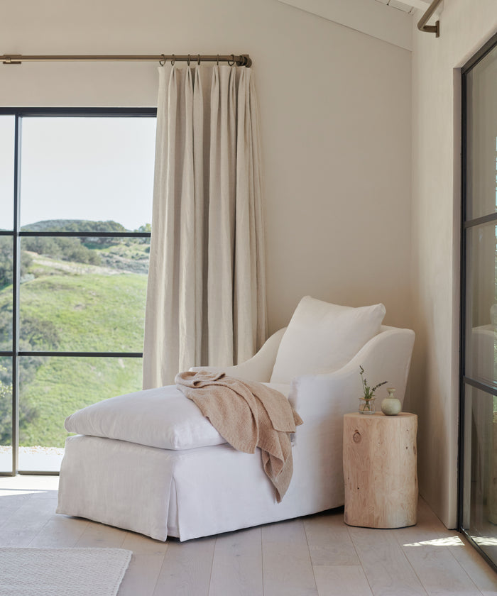 A tranquil spot with a white chaise lounge, beige throw, and light wood stump table is just one of many serene spaces accessed by JKH Membership, featuring soft curtains and hillside views through expansive windows.