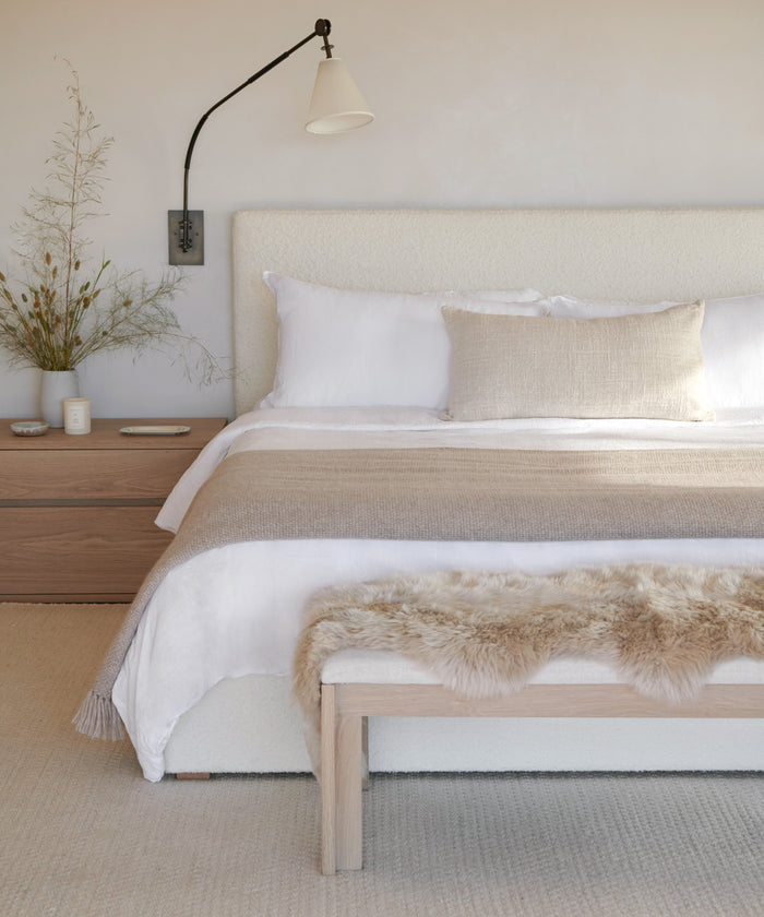 A minimalist bedroom features a white bed, beige throw blanket, and fur accent on a wooden bench. The JKH Membership touch is evident in the curated nightstand décor: vase with dried branches, candle, cup, and wall-mounted lamp.