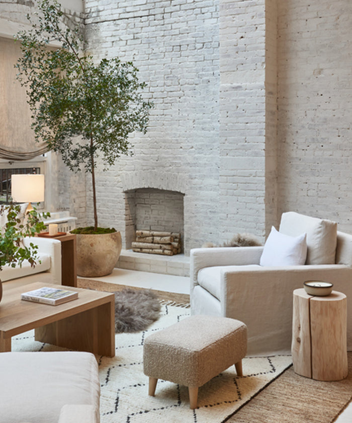 A cozy living room features white brick walls, potted tree, white armchairs, a wooden coffee table, and a decorative fireplace with stacked logs. JKH Membership-inspired décor in neutral tones enhances the calming atmosphere.