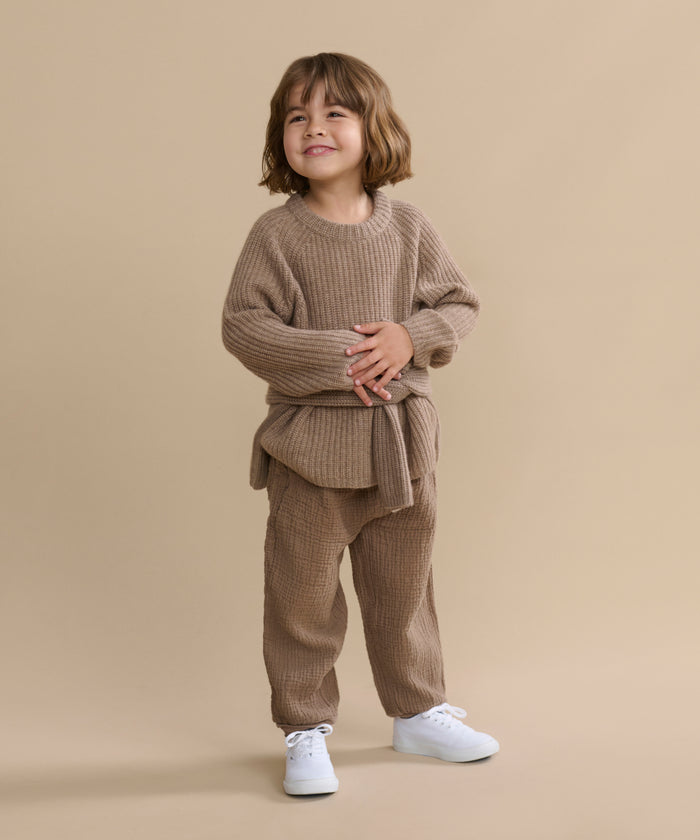 A young child with short brown hair smiles, wearing the cozy undyed stone Kids' Cashmere Fisherman sweater and matching pants with white sneakers, standing against a plain beige background.