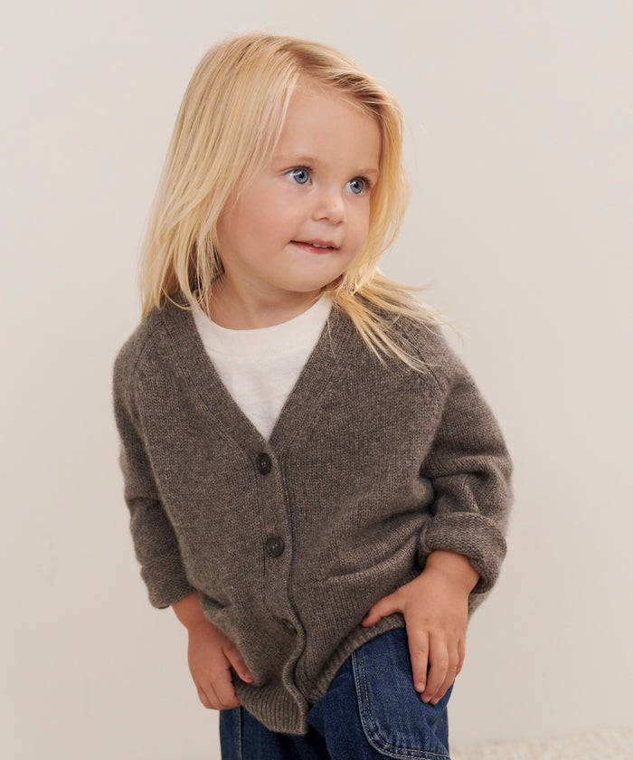 A young child with long blonde hair wears the Kids Cashmere Stanford Cardigan over a white shirt and blue jeans, standing and smiling slightly against a neutral background.