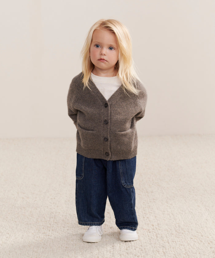 A young child with long blonde hair wears the Kids Cashmere Stanford Cardigan over a white shirt, loose blue jeans, and white shoes while standing on a beige carpet with hands behind their back, gazing slightly to the side.