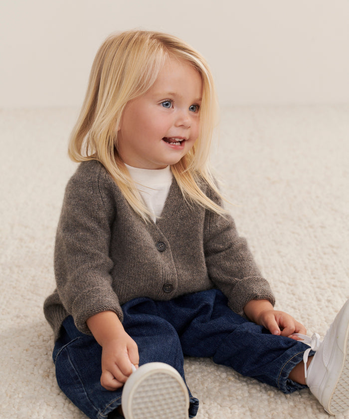 A young child with long blonde hair sits on a textured light carpet, smiling and looking off to the side while wearing the Kids Cashmere Stanford Cardigan over a white shirt, blue jeans, and white sneakers.