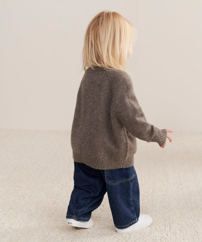A young child with blonde hair, wearing the Kids Cashmere Stanford Cardigan, blue jeans, and white shoes, walks on a light-colored carpet in a minimalist space, facing away from the camera.