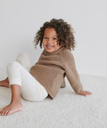 A young child with curly hair sits on a white carpet, smiling, wearing a cozy brown Kids Cotton Fisherman sweater and white ribbed leggings.